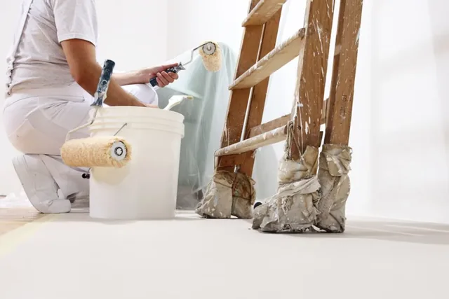 A painter on a ladder applying paint to an interior wall with a brush and roller